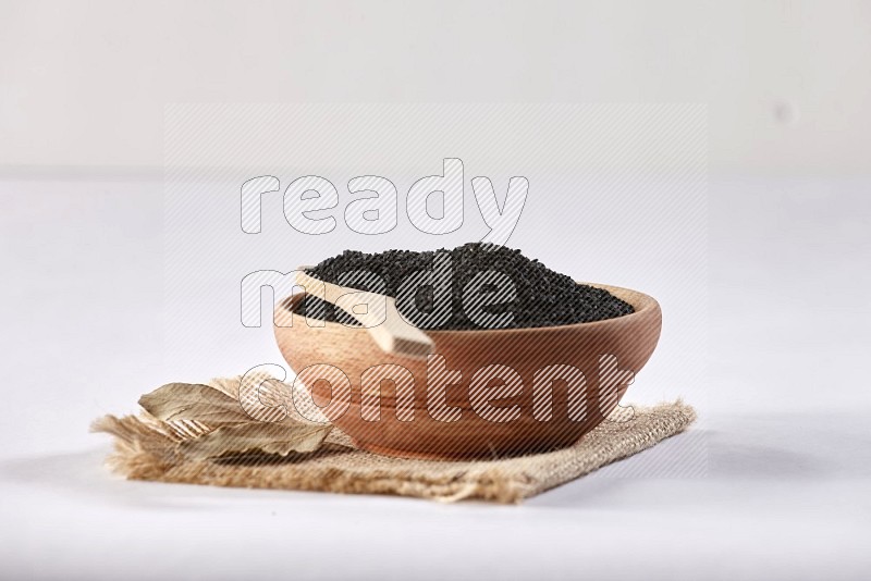 A wooden bowl and wooden spoon full of black seeds on a piece of burlap on a white flooring