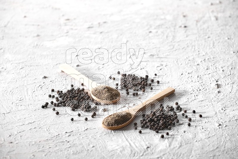 2 wooden spoons full of black pepper powder and black pepper beads spread on textured white background