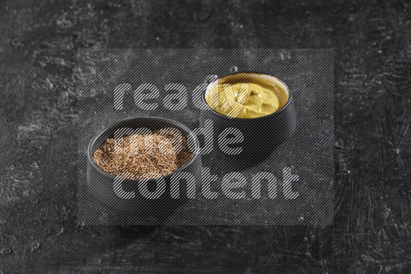 2 black pottery bowls full of mustard seeds and mustard paste on black flooring