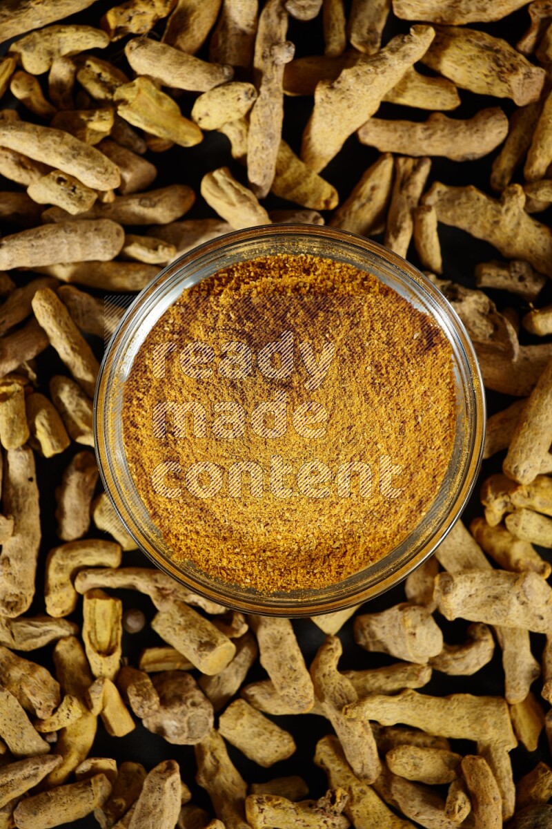 وعاء زجاجي ممتلئ ببودرة الكركم وأصابع الكركم المجففة بجانبه على خلفية سوداء