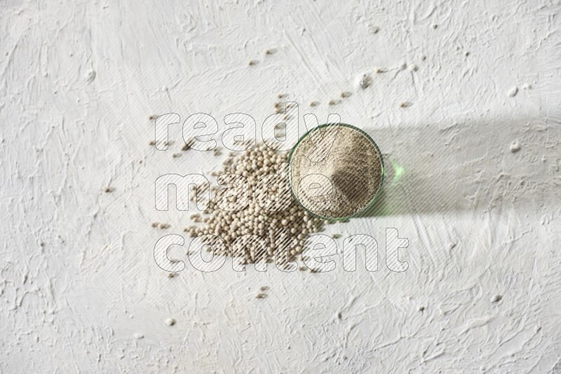 A green glass cup full of white pepper powder with white pepper beads on textured white flooring