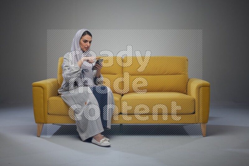 A Saudi woman wearing a light gray Abaya and white head scarf sitting on a yellow sofa and using her phone eye level on a grey background