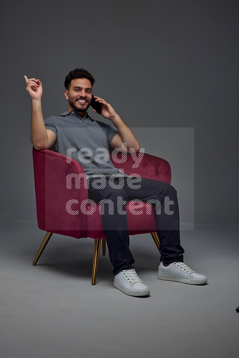 A man wearing casual talking in the phone and making multi hand gestures while sitting on a burgundy chair different angles eye level on a gray background