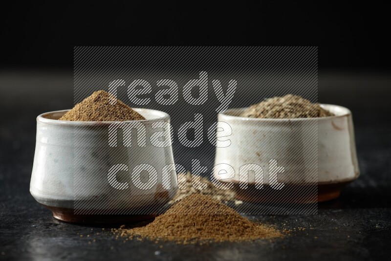 2 beige bowls, one full of cumin powder and the other full of seeds on a textured black flooring