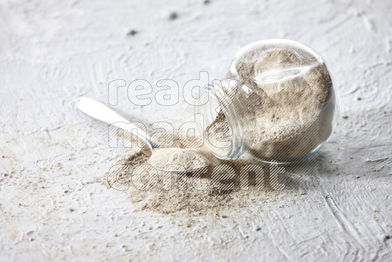 A flipped herbal glass jar and metal spoon full of white pepper powder with spilled powder on textured white flooring