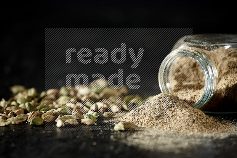 A flipped glass spice jar full of cardamom powder and powder spilled out of it with cardamom seeds spreaded on textured black flooring