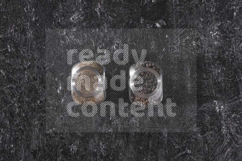 2 glass spice jars full of black pepper powder and black pepper beads on textured black flooring