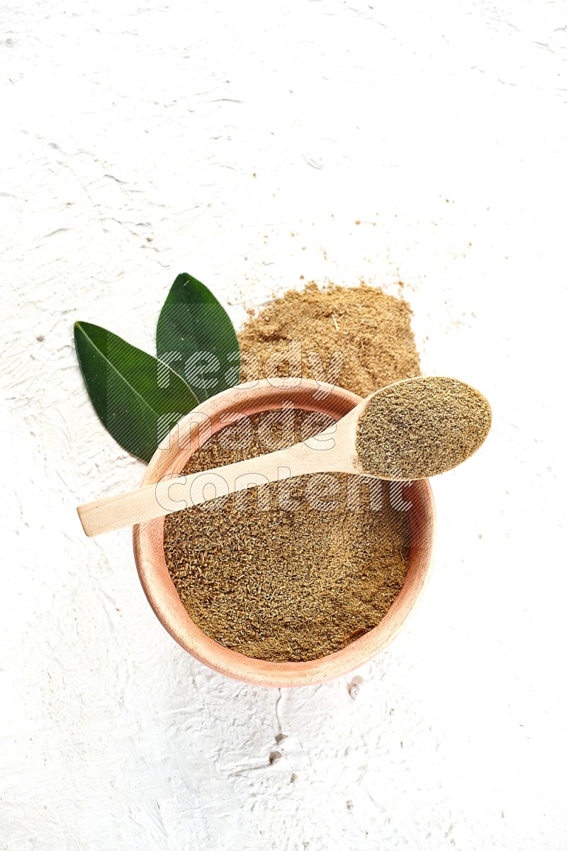 A wooden bowl and wooden spoon full of cumin powder on textured white flooring