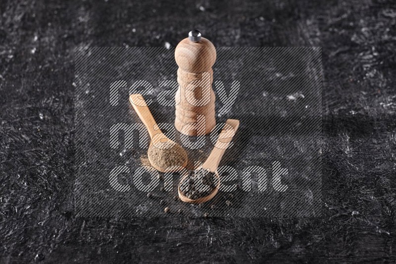 Black pepper powder and black pepper beads in wooden spoons and wooden grinder on a textured black flooring