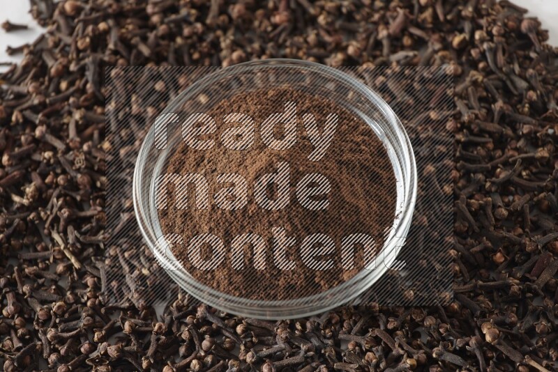 A glass bowl full of cloves powder and cloves grains spread on white flooring
