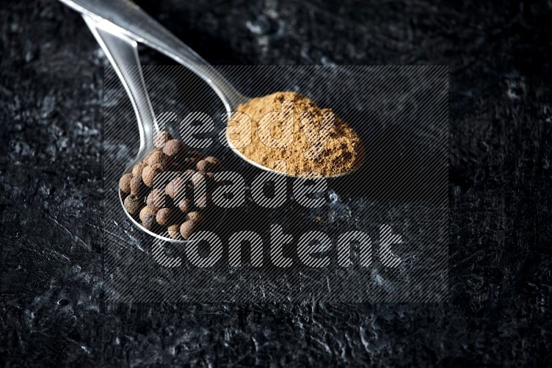 2 metal spoons full of allspice powder and whole balls on a textured black flooring