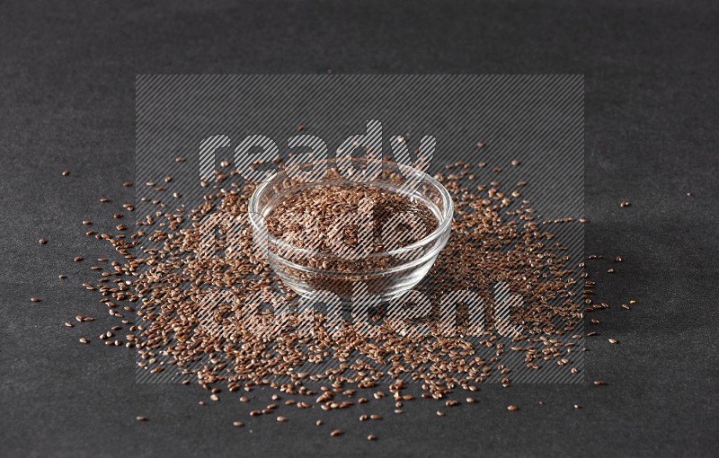 A glass bowl full of flaxseeds surrounded by the seeds on a black flooring