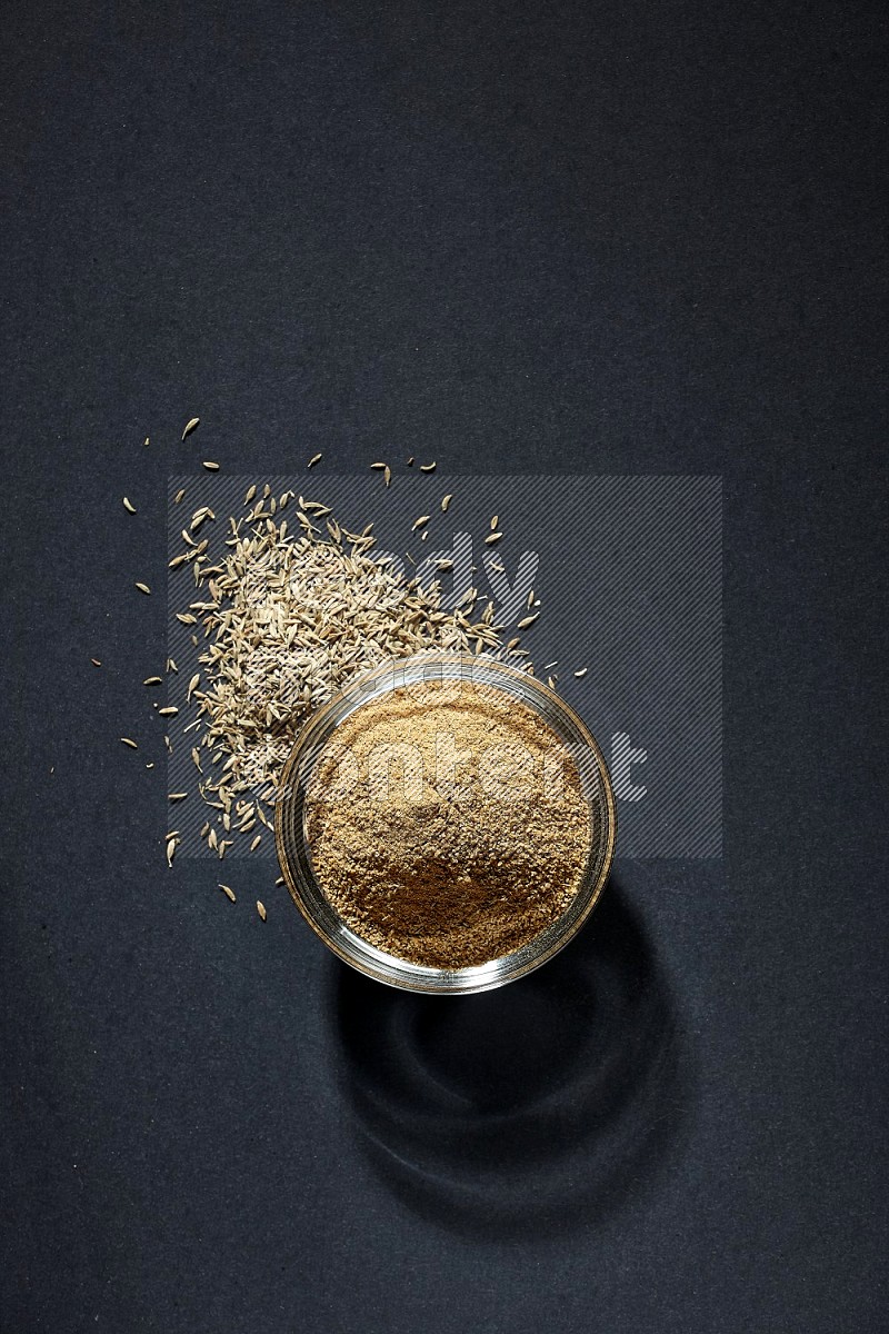 A glass bowl full of cumin powder with cumin seeds beside it on black flooring