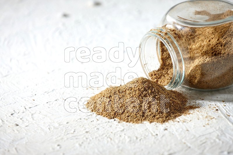 A flipped glass spice jar full of cumin powder and powder spilled out on textured white flooring