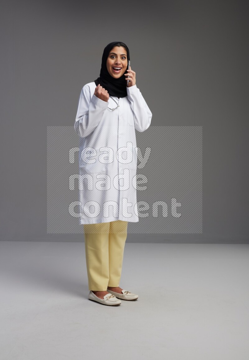 Saudi woman wearing lab coat with stethoscope standing talking on phone on Gray background