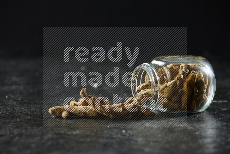 وعاء زجاجي للتوابل مقلوب وممتلئ بأصابع الكركم المجففة وبعض أصابع الكركم خارج الوعاء على خلفية سوداء