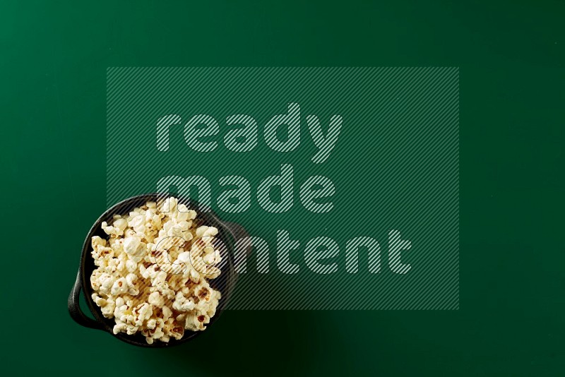 A black ceramic bowl full of popcorn on a green background in a top view shot