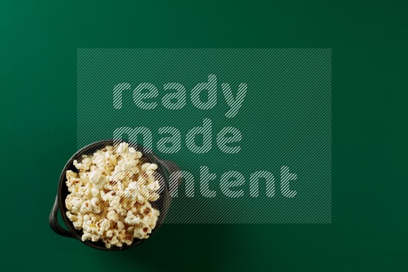 A black ceramic bowl full of popcorn on a green background in a top view shot