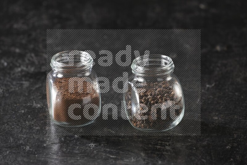 Glass spice jars full of cloves and cloves powder on textured black flooring
