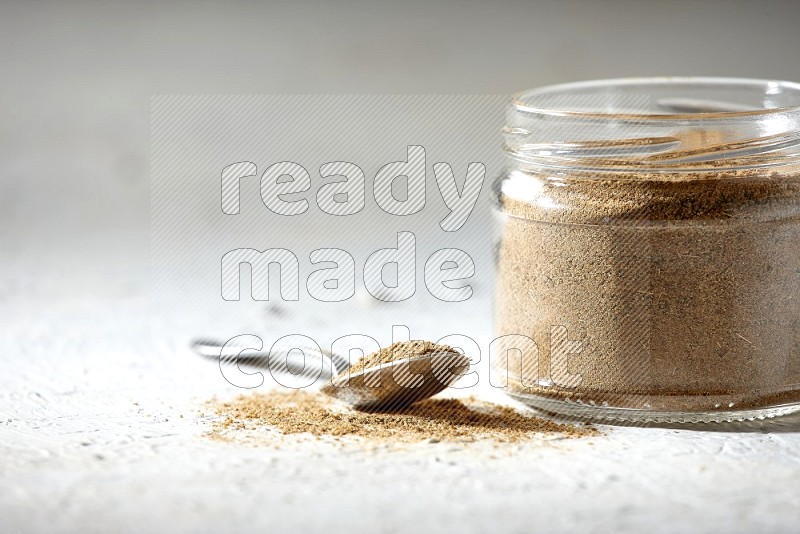 A glass jar and a metal spoon full of cumin powder on textured white flooring