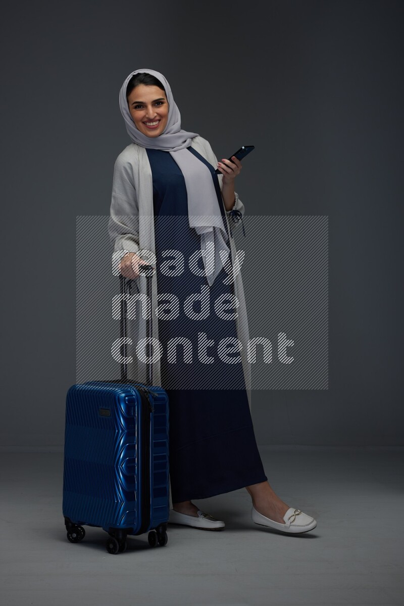 A Saudi woman wearing a light gray Abaya and head scarf standing holding a phone and being shocked  eye level on a grey background