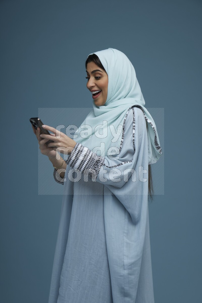 A Saudi woman Texting in a blue background wearing a blue Abaya with hijab
