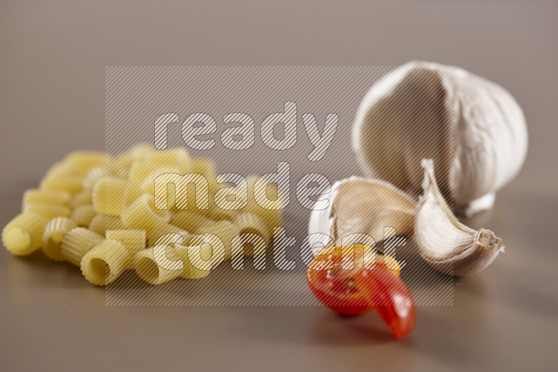 Raw pasta with different ingredients such as cherry tomatoes, garlic, onions, red chilis, black pepper, white pepper, bay laurel leaves, rosemary and cardamom on beige background