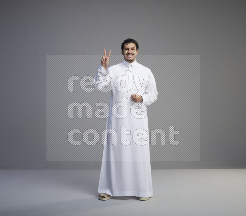 A Saudi man standing wearing thob with face painting interacting with the camera on gray background
