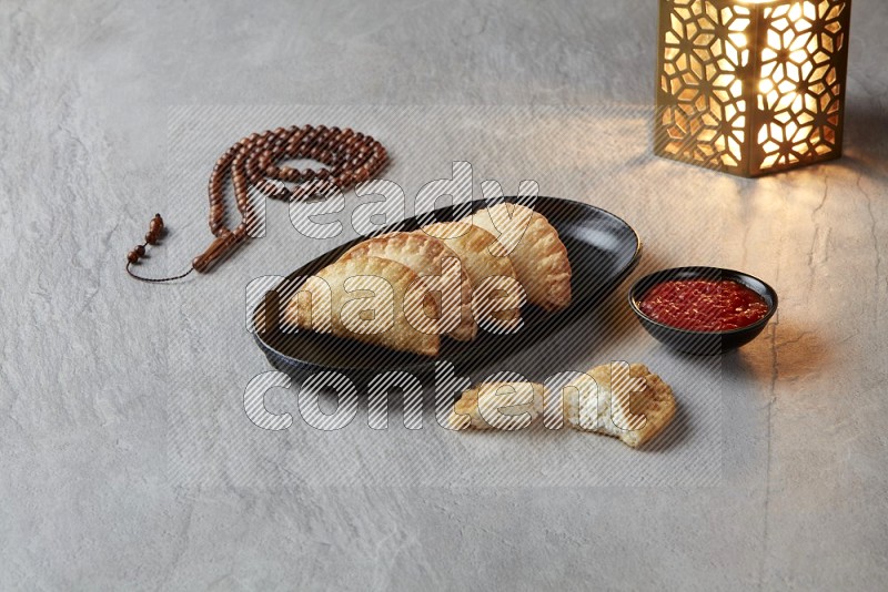 Four fried sambosas in an oval shaped black plate, beside a cut cheese sambosa, a brown misbaha and a golden lantern on a gray background