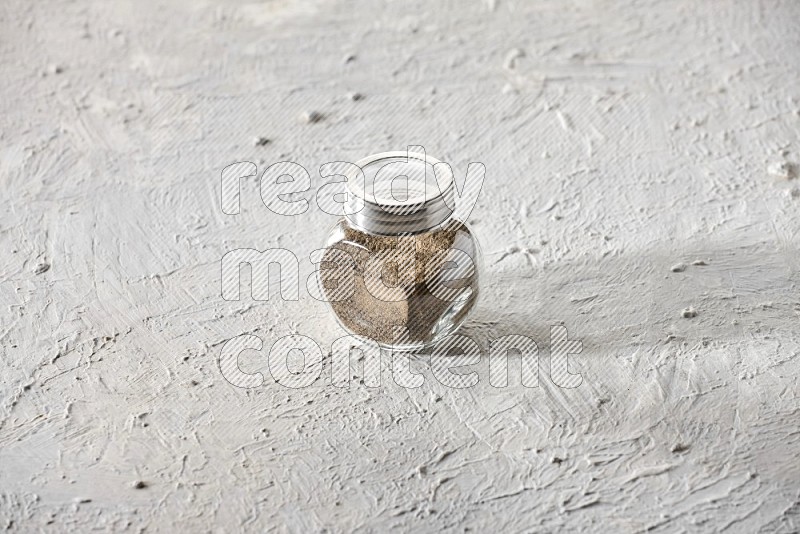 A glass spice jar full of powder black pepper on textured white flooring