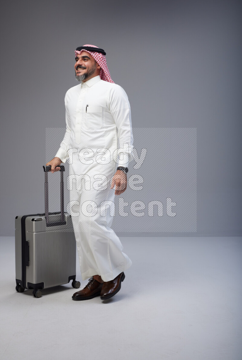Saudi man wearing Thob and red Shomag standing holding Travel bag on Gray background