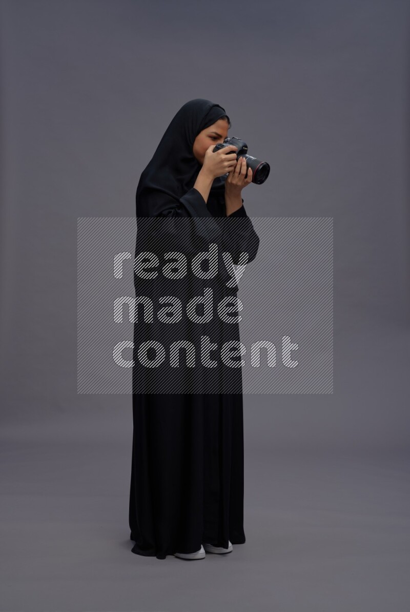 Saudi woman wearing Abaya standing holding Camera on gray background