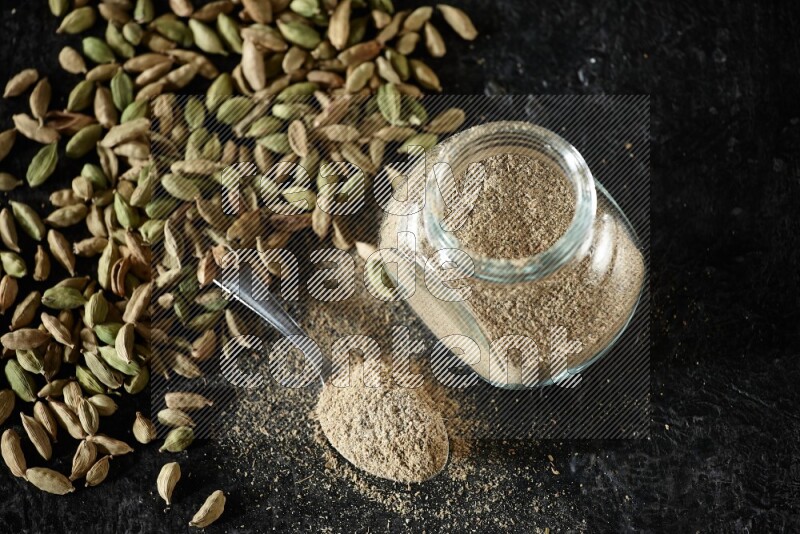 A glass spice jar and a metal spoon full of cardamom powder and cardamom seeds spreaded on textured black flooring