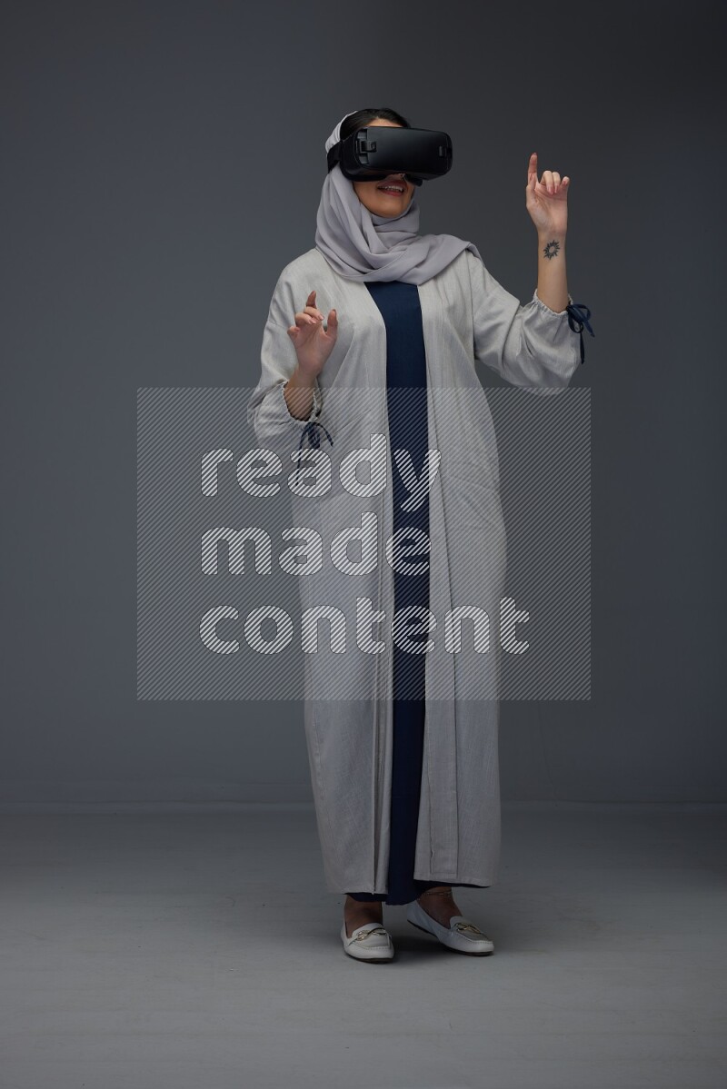 A Saudi woman wearing a light gray Abaya and head scarf standing and wearing VR eye level on a grey background