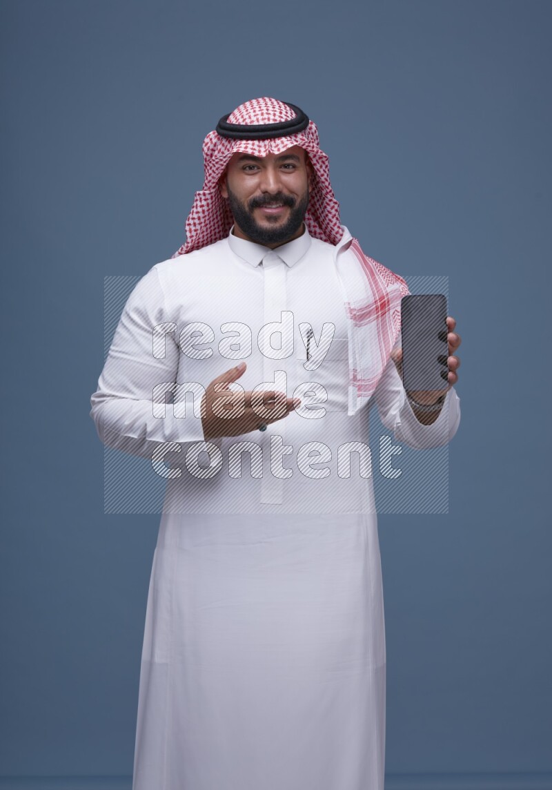 A man Showing his smartphone Screen on Blue Background wearing Saudi Thob