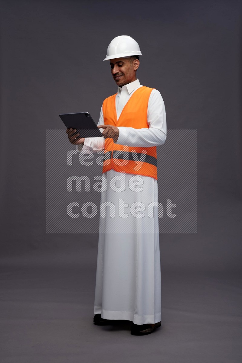 Saudi man wearing thob with engineer vest standing working on tablet on gray background