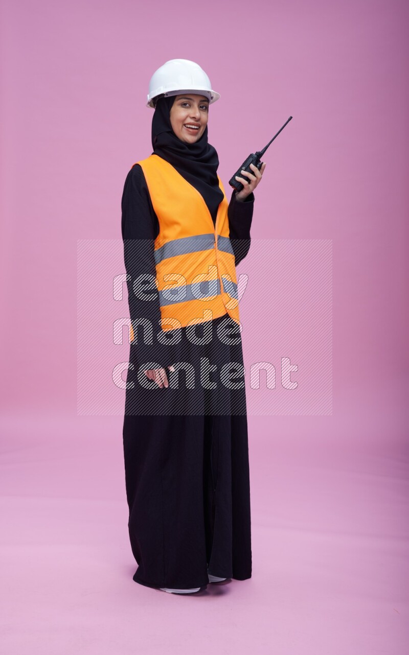 Saudi woman wearing Abaya with engineer vest and helmet standing holding walkie-talkie on pink background