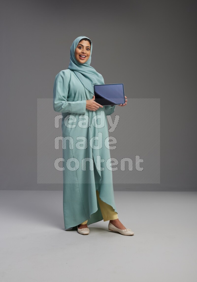 Saudi Woman wearing Abaya standing showing tablet to camera on Gray background