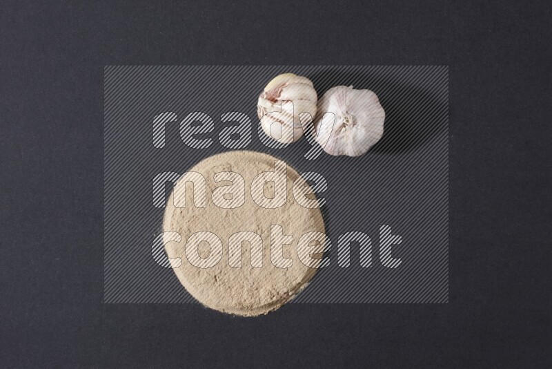 Garlic powder in a circle shape and beside it 2 garlic bulbs on a black flooring
