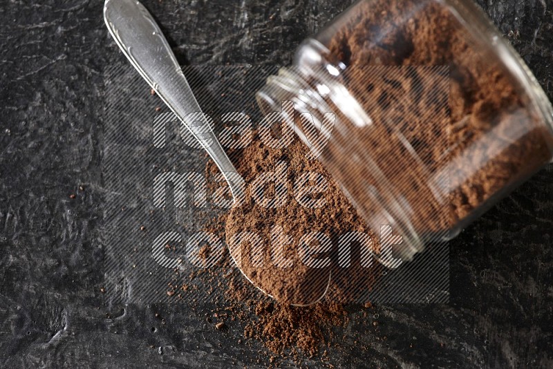 A flipped glass jar and metal spoon full of cloves powder with cloves spread on a textured black flooring