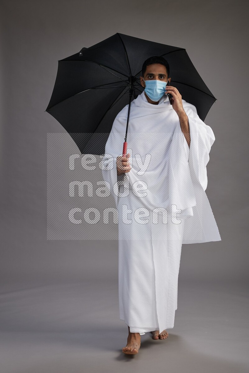 A man wearing Ehram and mask Standing holding umbrella on gray background on gray background