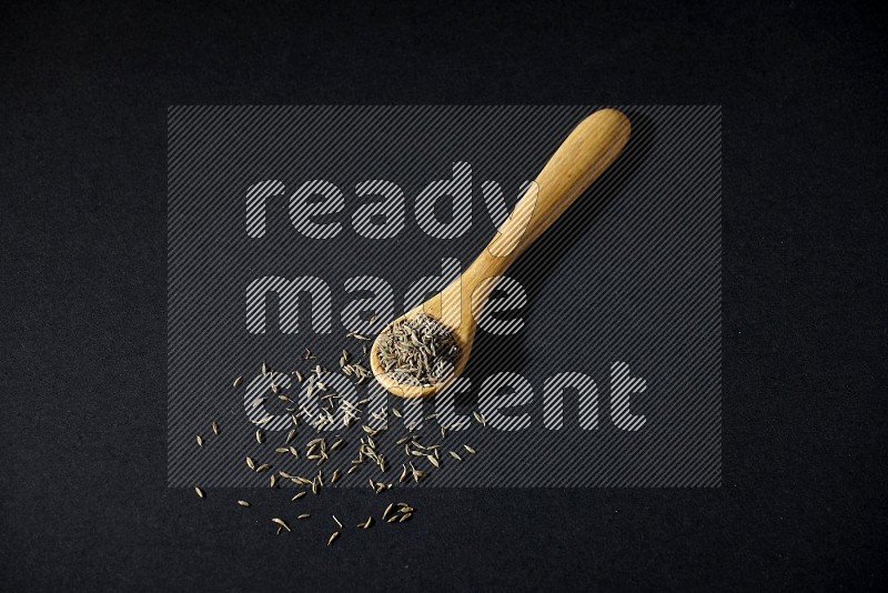 A wooden spoon full of cumin seeds on black flooring