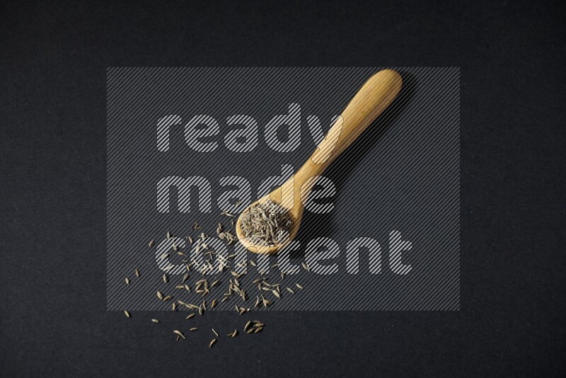 A wooden spoon full of cumin seeds on black flooring