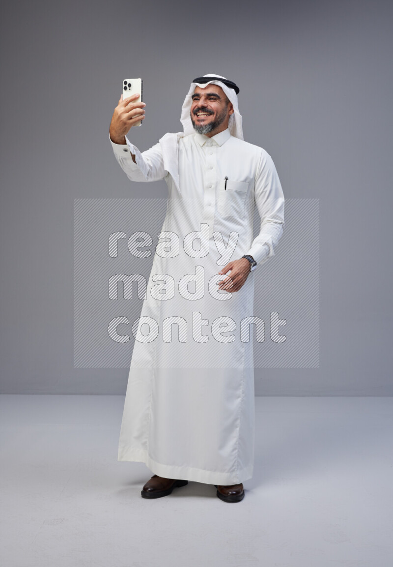 Saudi man Wearing Thob and white Shomag standing taking selfie on Gray background