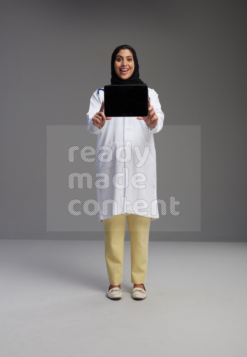 Saudi woman wearing lab coat with stethoscope standing showing tablet to camera with sign in the back on Gray background