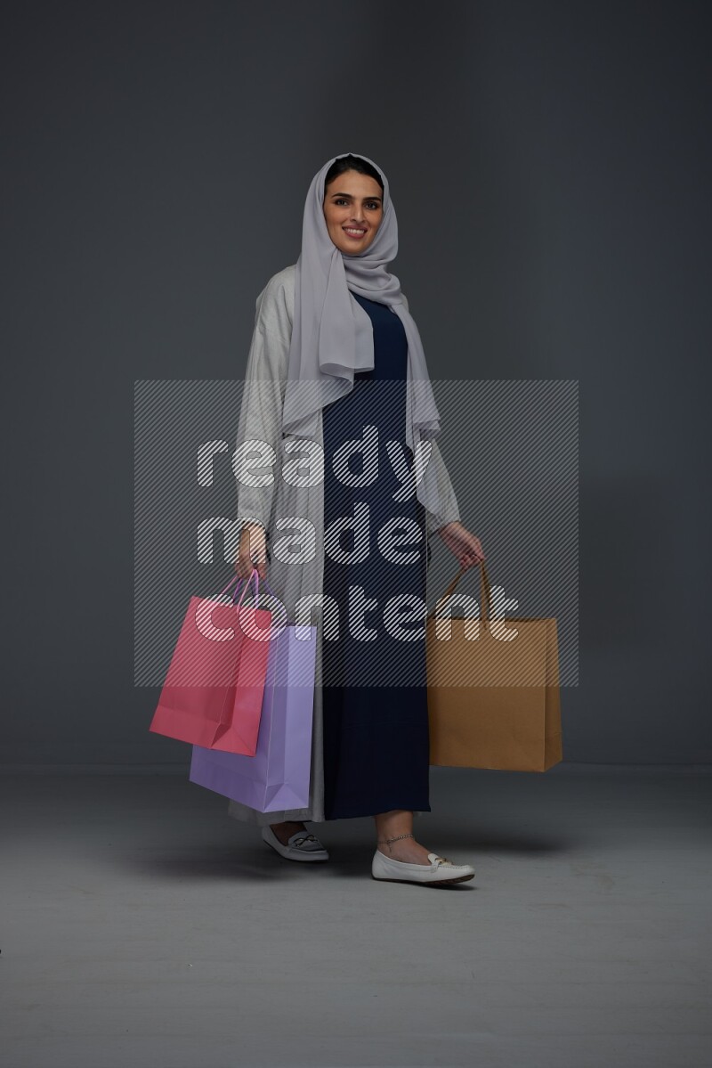 A Saudi woman wearing a light gray Abaya and head scarf standing and holding shopping bags on a grey background