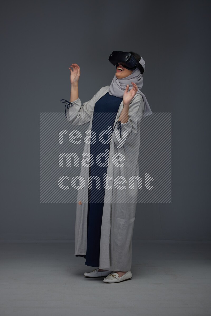 A Saudi woman wearing a light gray Abaya and head scarf standing and wearing VR eye level on a grey background