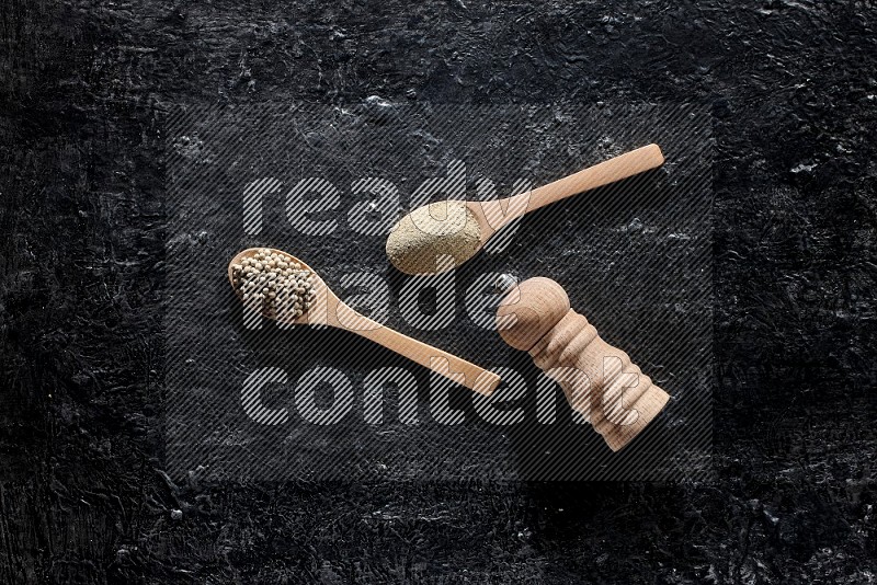 2 wooden spoons one full of white pepper powder and the other with pepper beads and a wooden pepper mill on textured black flooring