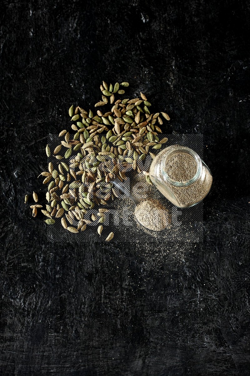 A glass spice jar and a metal spoon full of cardamom powder and cardamom seeds spreaded on textured black flooring