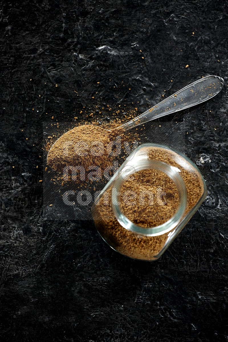 A glass spice jar and a metal spoon full of cumin powder on a textured black flooring